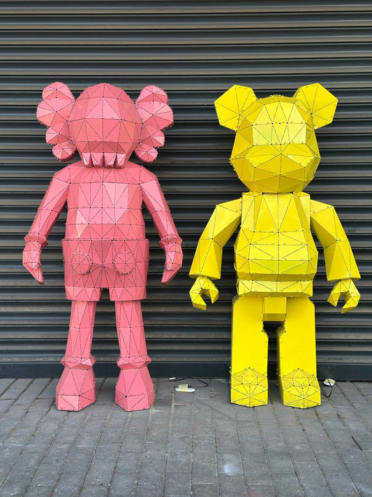 Two large, low-poly metal bear sculptures in pink and yellow against a grey shutter wall.