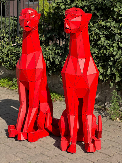 Two red geometric metal sculptures of sitting big cats against a green hedge.