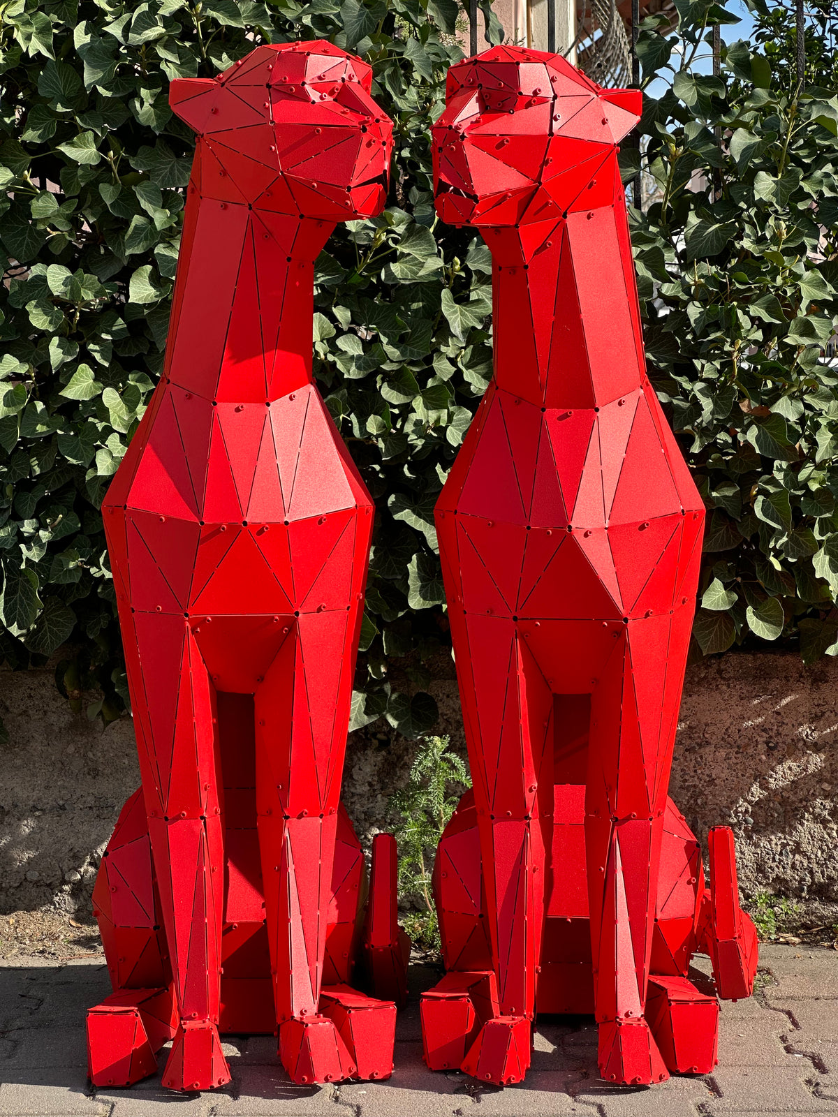 Two red geometric metal sculptures of sitting cheetahs facing each other against green foliage