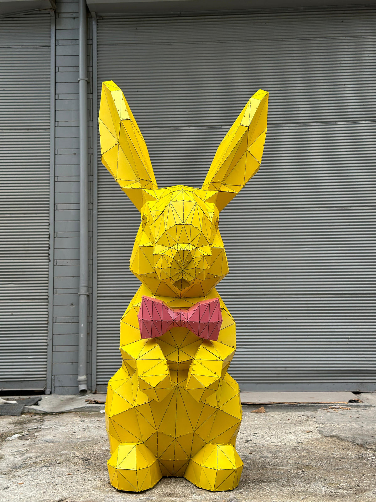 Geometric yellow metal rabbit sculpture wearing a pink bow tie against a grey shutter background